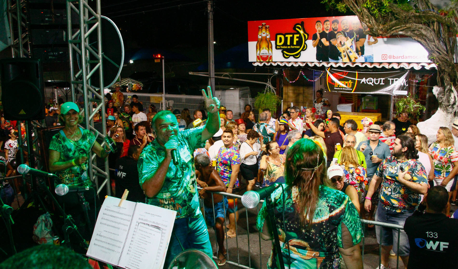 pessoas na rua em frente a palco com banda tocando e bares ao fundo